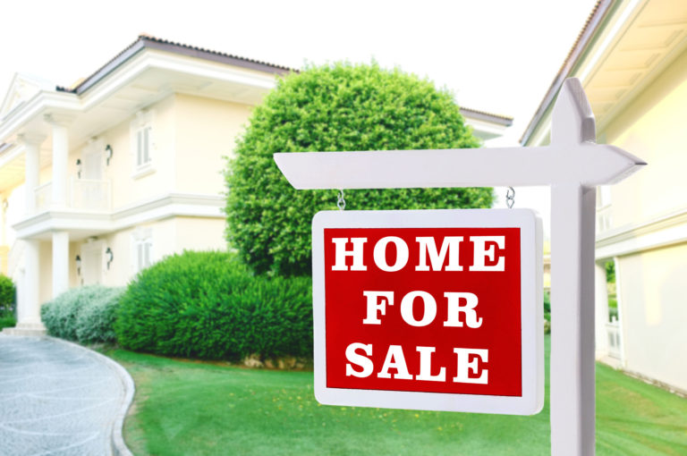 'Home For Sale' sign in front of a yellow house with shrubs.