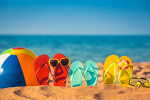 Three pairs of flipflops, sunglasses, and a beachball sit on the beach.